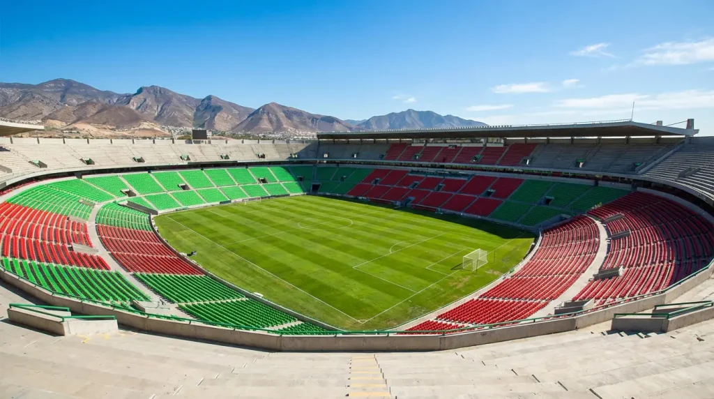 Estadio de fútbol mexicano con gradas coloridas y montañas al fondo bajo cielo azul